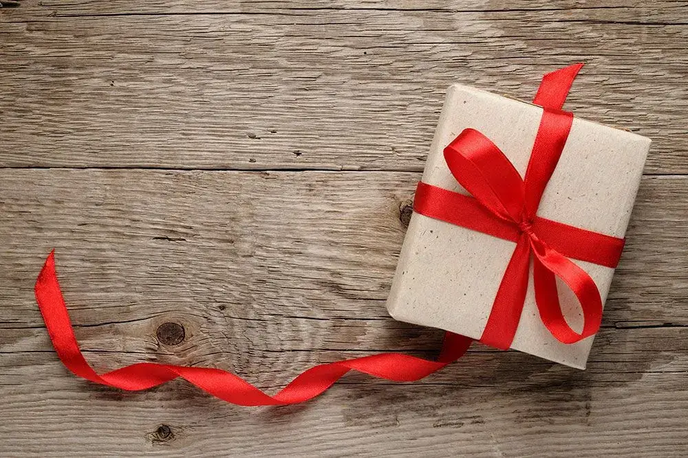 A holiday gift box, wrapped in light paper with a bright red ribbon and bow, rests on a rustic wooden surface