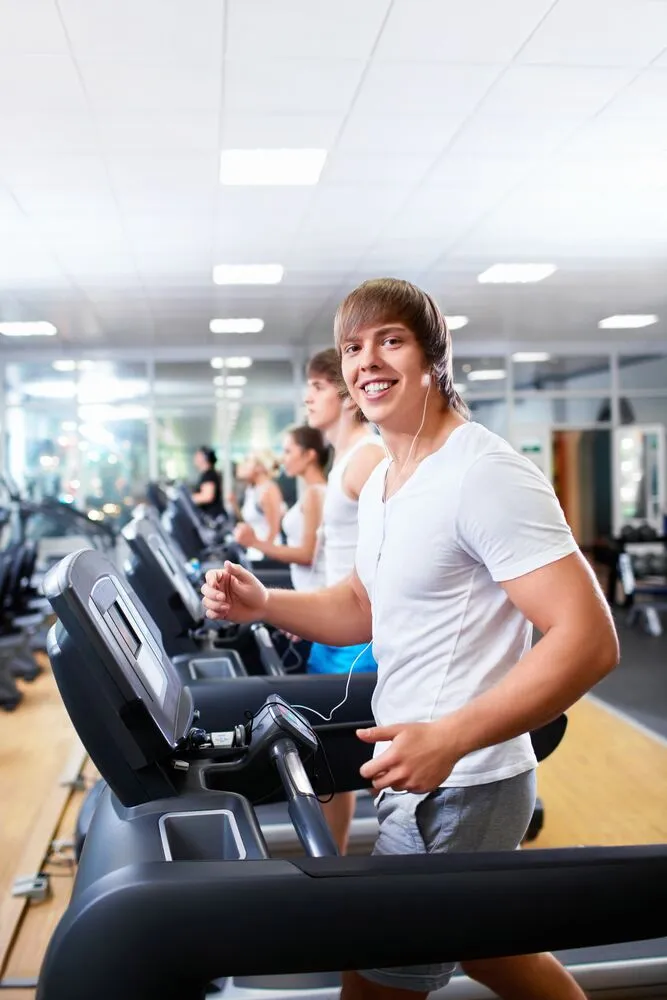 A happy young man in a white shirt and earbuds running on a treadmill at the gym