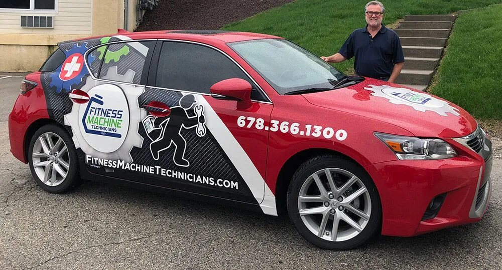  Ed Bauer stands proudly beside a red car wrapped with "Fitness Machine Technicians" branding