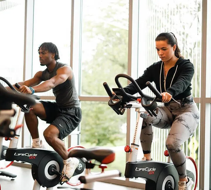 A man and a woman actively riding stationary bikes in a modern, brightly lit gym with large windows