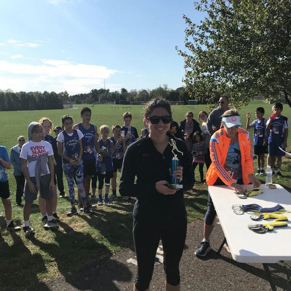 A woman proudly holds a trophy at a 5K race awards ceremony with children and adults gathered
